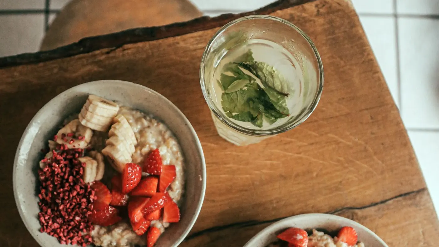 Dos tazones de avena con banano y fresas sobre una mesa de madera, vistos desde arriba, junto a un vaso de café frío y otro de agua con hierbabuena