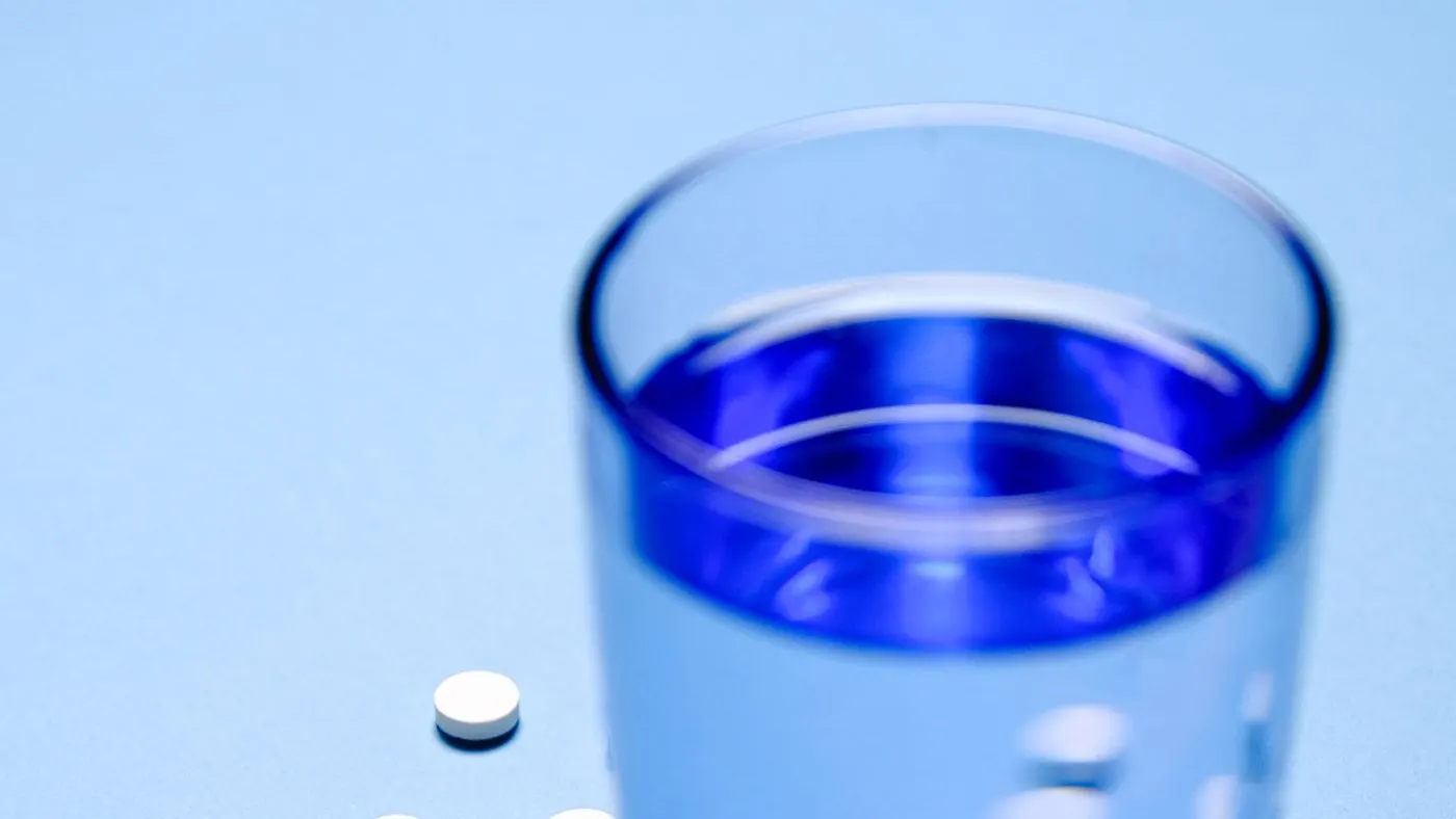 Vaso de agua y cuatro pastillas blancas sobre una superficie azul, imagen para la guía del enalapril