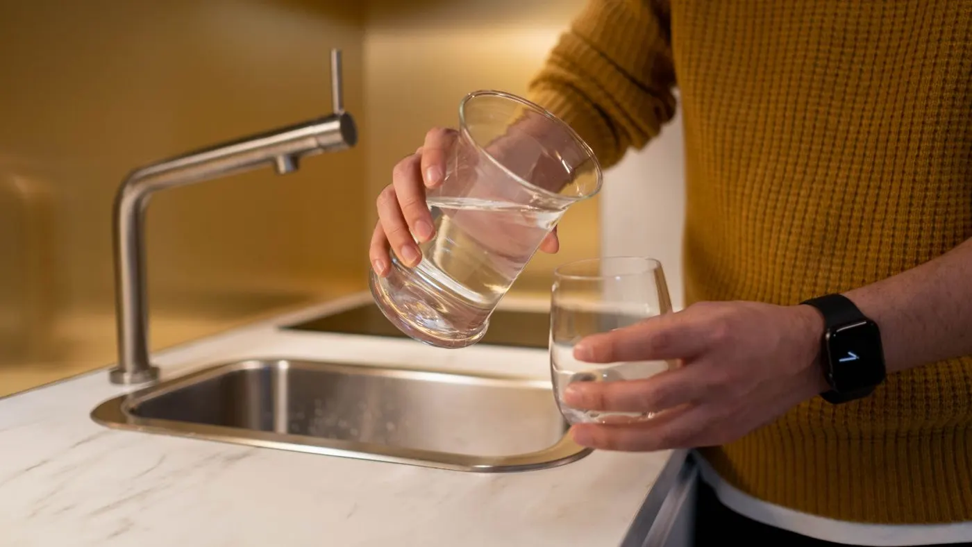 Persona con suéter mostaza que vierte agua de un vaso a otro junto al lavaplatos de la cocina, imagen para la guía del irbesartán