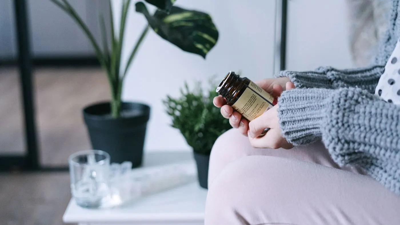 Manos con suéter de lana gris que sostienen un frasco de vidrio oscuro con etiqueta, junto a una mesa blanca con un vaso de agua y un pastillero, entre plantas de interior
